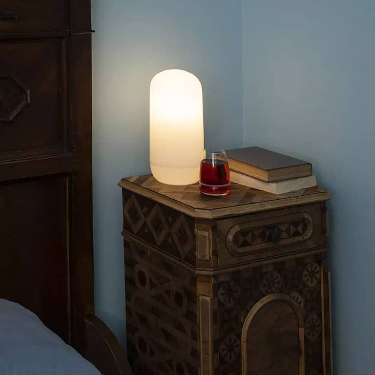 Wooden carved bedside table with a glowing white lamp, a glass of red beverage, and two stacked books against a blue wall.