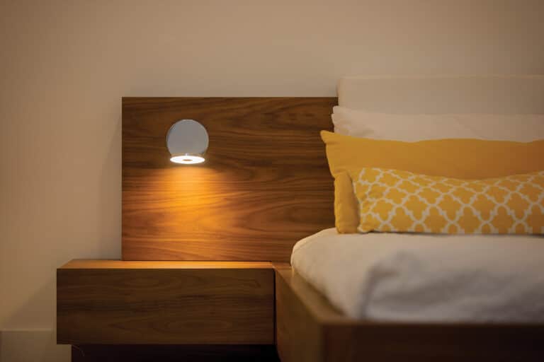 Wooden headboard with a small nightstand and a round wall lamp casting warm light over a neatly made bed with yellow pillows.