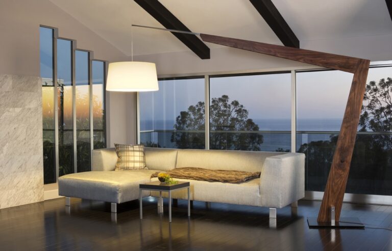 Modern living room with an L-shaped gray sofa, a floor lamp, and a wooden sculptural beam framing a sea-view through large glass doors.