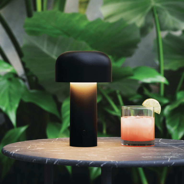 Black mushroom-shaped lamp glows on a round marble table next to a pink cocktail with a lime wedge, with lush green plants in the background.