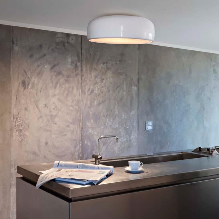 Stainless steel kitchen counter with sink, newspaper and cup, concrete-panel wall, and a circular white ceiling light.