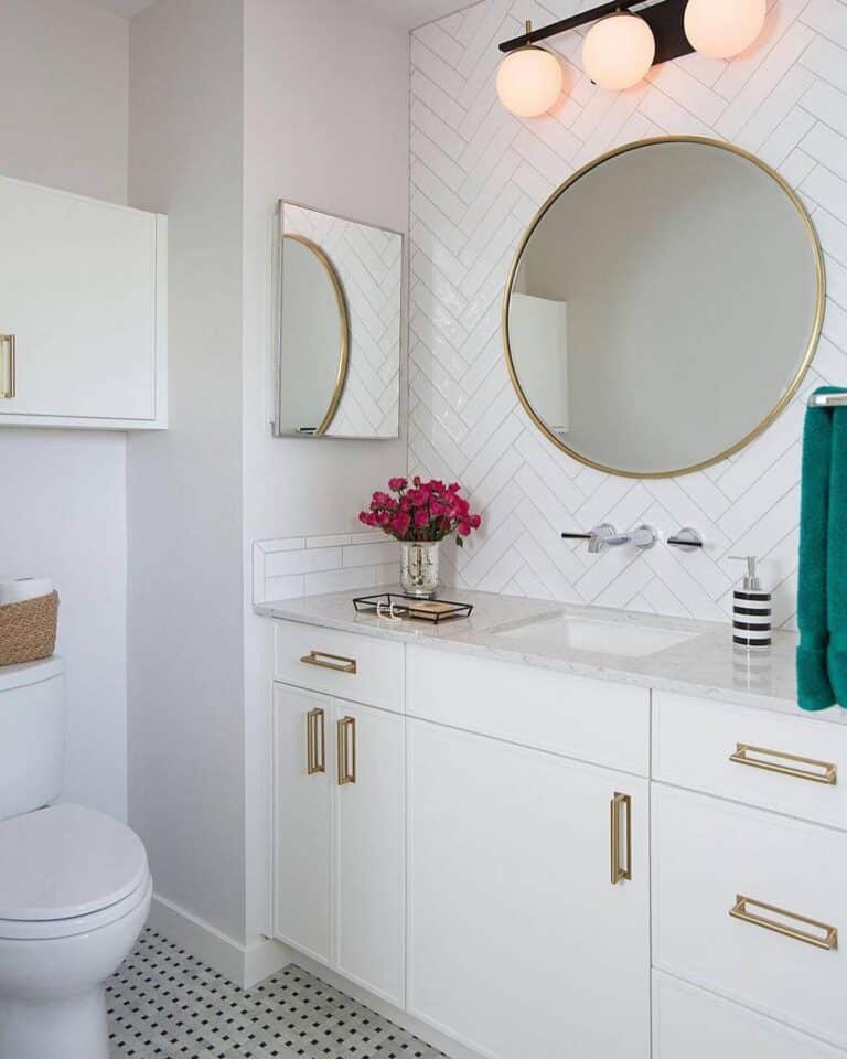 Modern white bathroom with a double vanity, round gold-framed mirror, herringbone tiled wall, and pink flowers on the counter.