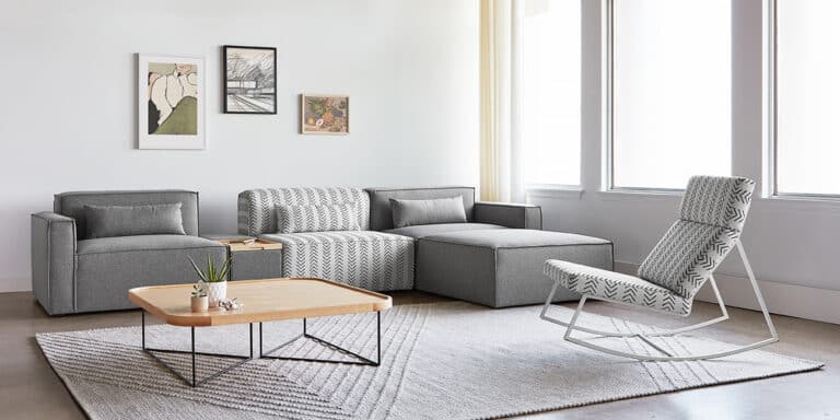 Bright living room with a gray sectional sofa, patterned pillows, and a wooden coffee table by large windows.  A white rocking chair sits on a light rug beside the seating area.