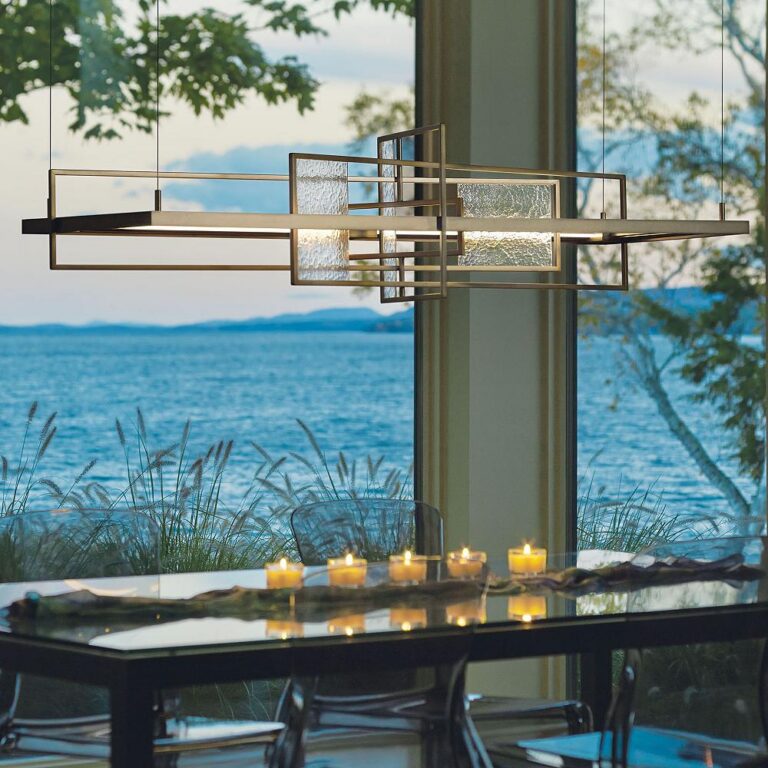Modern geometric chandelier above a glass dining table by a large window overlooking a lake/ocean with candles lit along the table edge.