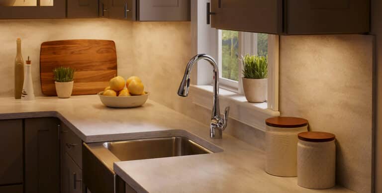 Modern kitchen counter with a shiny chrome faucet, sink, and a bowl of lemons under warm under-cabinet lighting by a window. A wooden cutting board, oil bottle, and potted plants sit along the counter.