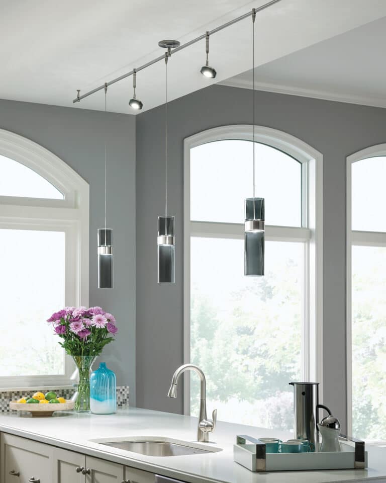 Modern kitchen island with three pendant lights, gray walls, arched windows, and a sink with a curved faucet nearby a vase of pink flowers and citrus tray.