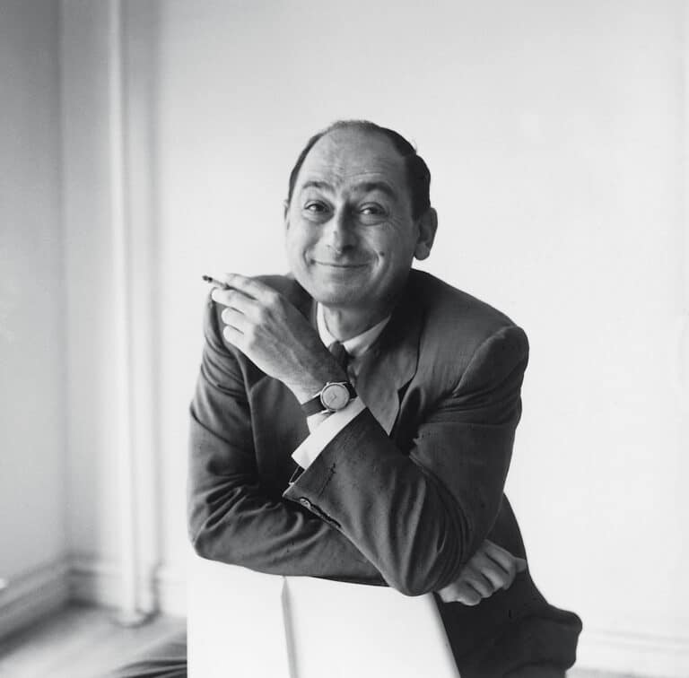Smiling man in a suit, leaning on a white surface while holding a cigarette.