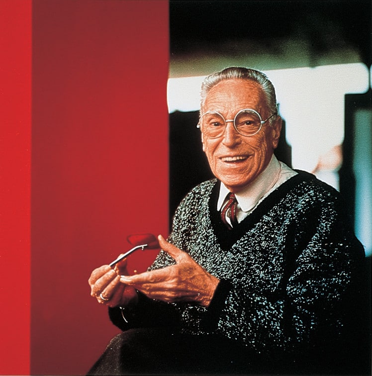 Smiling elderly man with round glasses, wearing a sparkly black sweater, holding his eyeglasses.