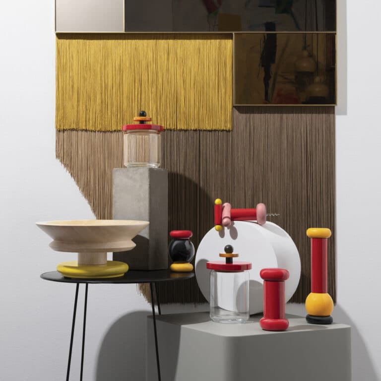 Colorful wooden toys and glass jars arranged on a table and pedestal against a textured yarn wall in a gallery setting
