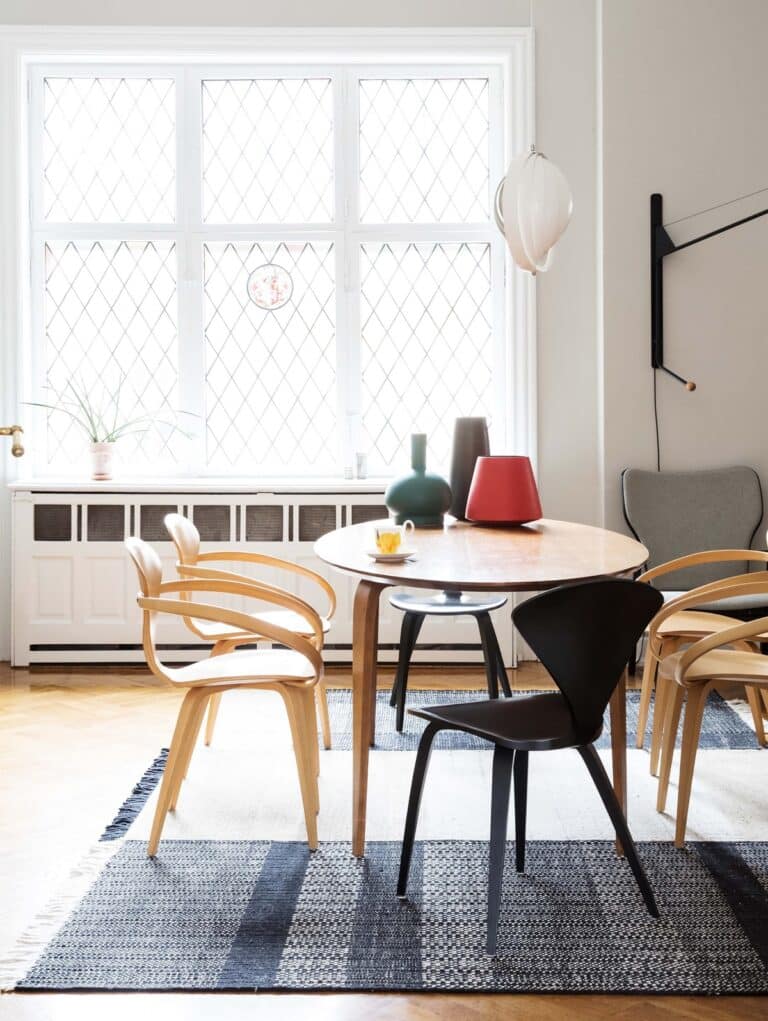 Bright dining area with a round wooden table, mixed light-wood and black chairs, and a large lattice-window letting in light.