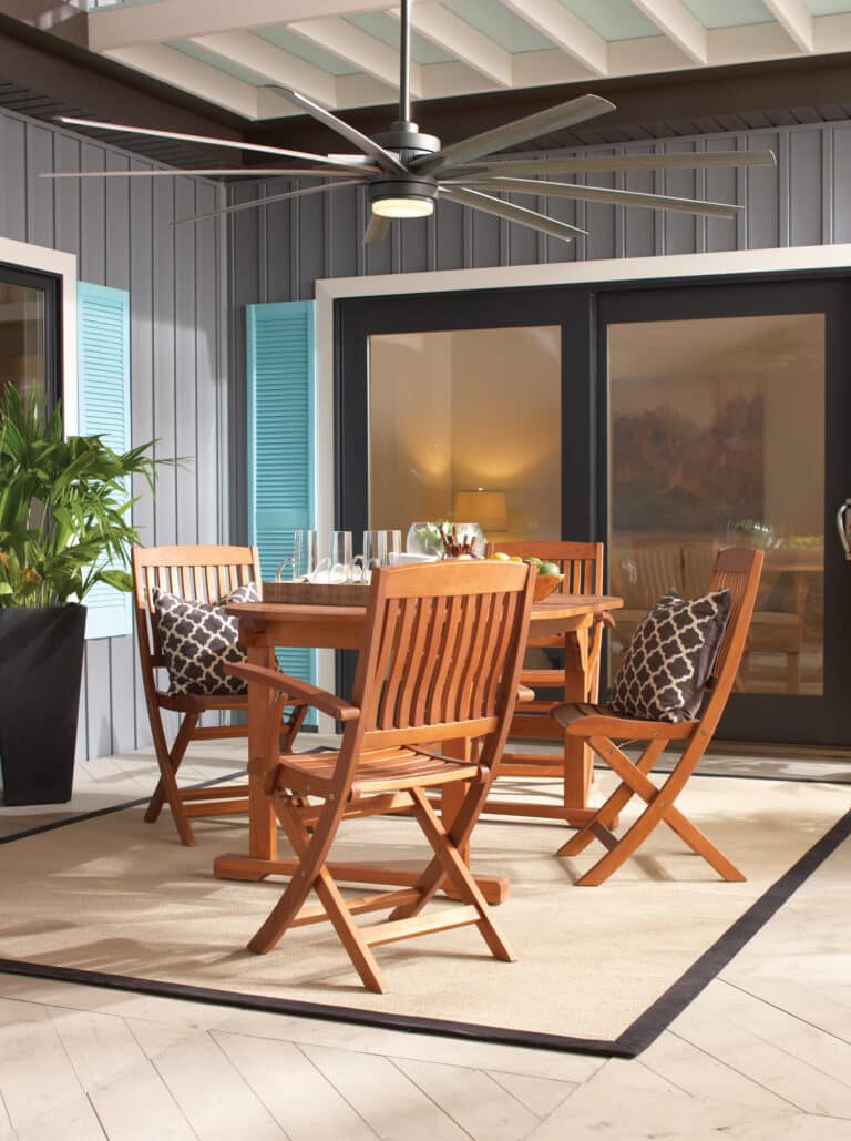 Outdoor dining area with wooden table and chairs on a patio under a large ceiling fan with a frosted light fixture