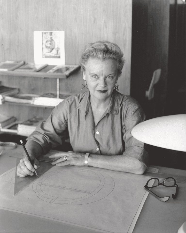 Older woman seated at a drafting table, drawing circular patterns with a pencil; glasses and a lamp sit nearby.