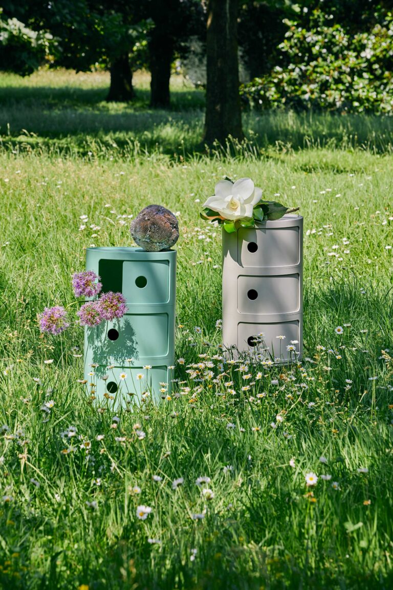 Pastel green and gray stacked storage chests in a sunlit meadow, topped with a white magnolia and green leaves amid daisies.