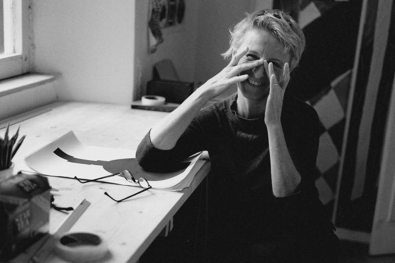 Smiling person at a cluttered desk, hands near their face with glasses and papers strewn about.