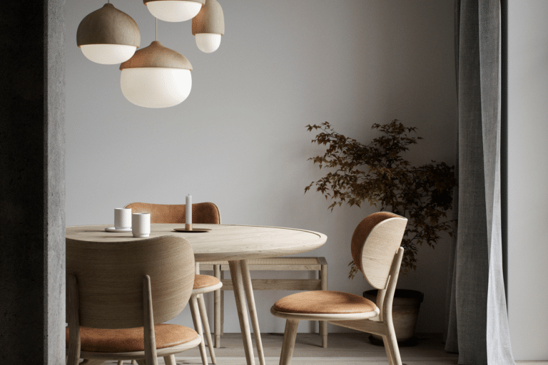 Round wooden dining table with matching chairs in a minimalist dining nook, lit by hanging pendant lights and a potted plant nearby.