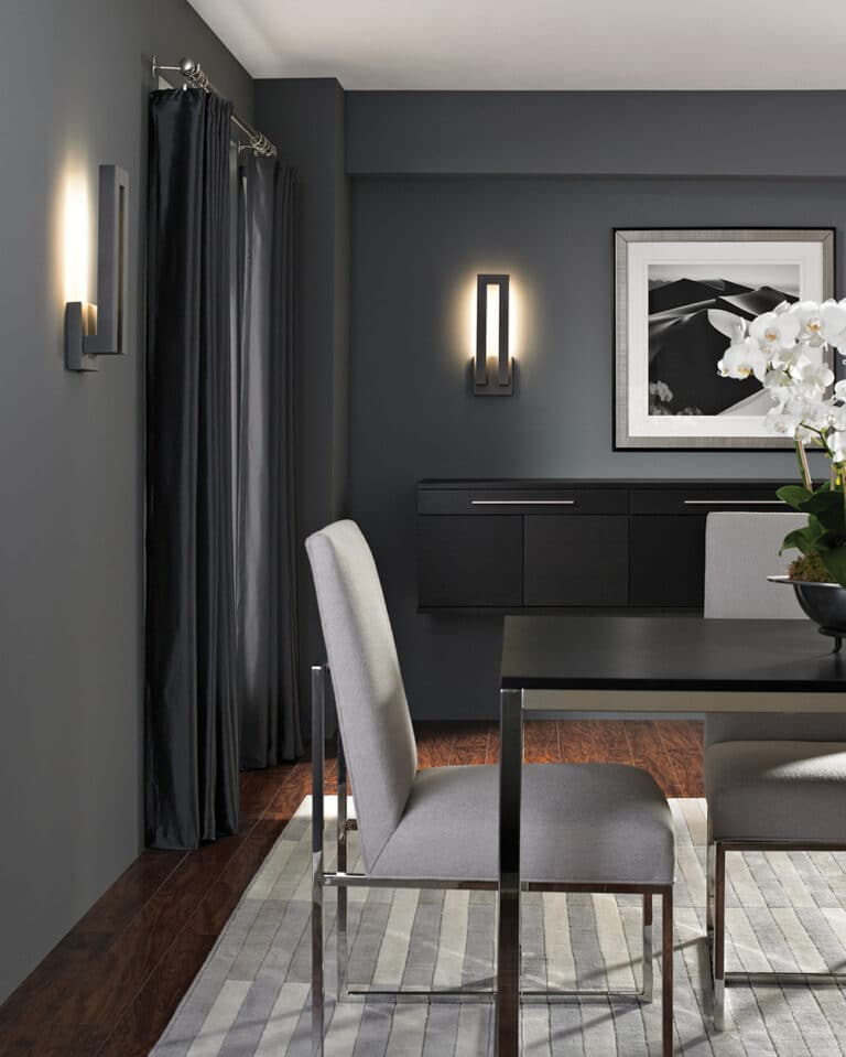 Modern dining room with dark gray walls, a black sideboard, and gray upholstered chairs around a black table with a white orchid centerpiece.