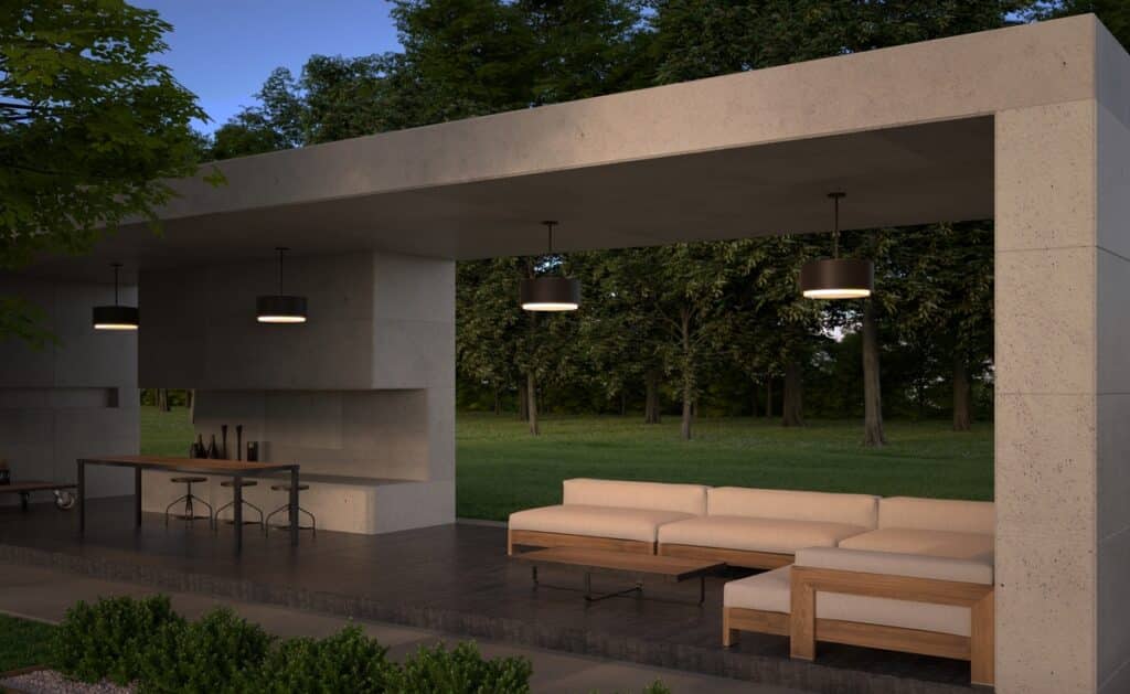 Open-air stone pavilion with beige sectional sofa, wooden coffee table, and a bar area with stools, set in a green park under hanging lights.