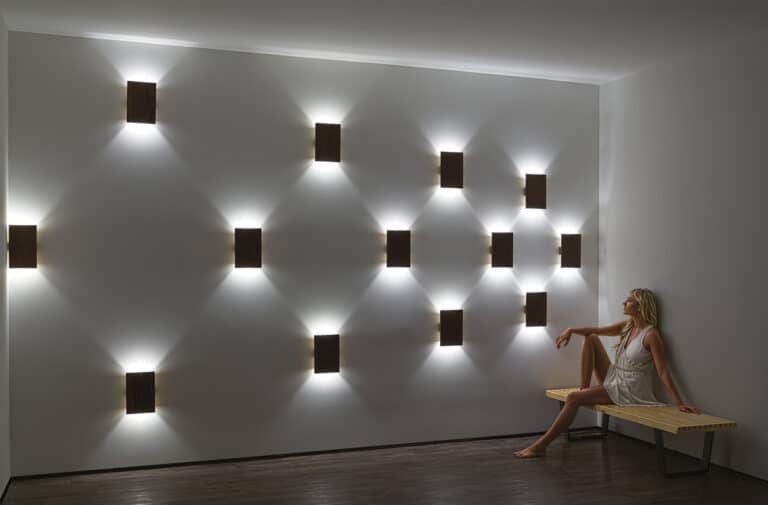 Minimalist room with square wall lights casting diamond-shaped glow; a woman sits on a wooden bench to the right.