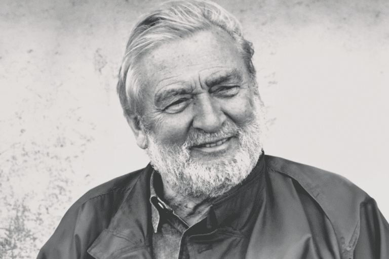 Smiling older man with a white beard and gray hair, wearing a dark jacket, against a textured light background.