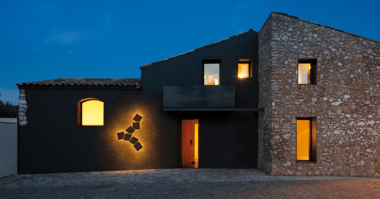 Night exterior of a modern house: dark stucco left wing and stone-clad right wing with warm light from windows and an open doorway.