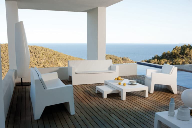 White outdoor seating area on a wooden deck overlooking the blue sea and distant hills, with a minimalist design.